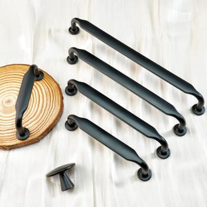 Könnte beinhalten: Eine Auswahl an schwarzen Schrankgriffen und einem Knauf, präsentiert auf einer weißen Oberfläche. Die Griffe in verschiedenen Größen haben ein gebogenes Design. Ein Griff ist auf einer Holzscheibe befestigt. Die Hardware ist aus Metall.