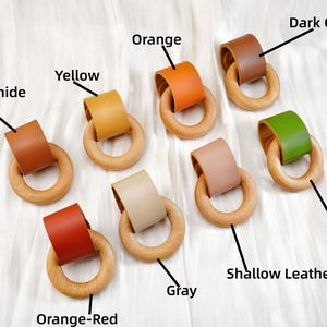 Peut inclure: Plusieurs anneaux de dentition en bois avec des accents de cuir de diff&eacute;rentes couleurs, notamment orange, jaune, vert et marron. Les anneaux sont dispos&eacute;s sur une surface blanche, pr&eacute;sentant les diff&eacute;rentes options de couleurs.