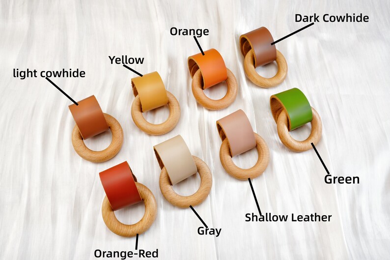 Peut inclure: Plusieurs anneaux de dentition en bois avec des accents de cuir de diff&eacute;rentes couleurs, notamment orange, jaune, vert et marron. Les anneaux sont dispos&eacute;s sur une surface blanche, pr&eacute;sentant les diff&eacute;rentes options de couleurs.