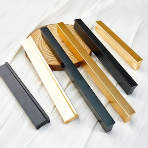 Può includere: Un assortimento di maniglie rettangolari per mobili in nero opaco, oro e crema. Le maniglie sono disposte su un tessuto bianco e una fetta di legno, mostrando le loro diverse finiture e design. Misure in centimetri.