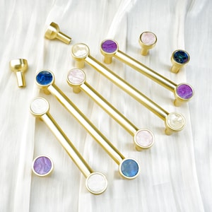 May include: A selection of gold-coloured cabinet pulls and knobs with colourful, gem-like accents. The pulls have a bar shape with circular ends in white, pink, blue, and purple. The knobs have a similar design. The hardware is arranged on a white, textured surface.
