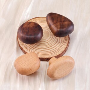 Może przedstawiać: Kolekcja drewnianych gałek do szafek w r&oacute;żnych kształtach i odcieniach drewna. Dwie ciemnobrązowe gałki leżą na drewnianym plastrze, a pod nimi dwie jaśniejsze gałki. Gałki są ułożone na kremowej tkaninie.