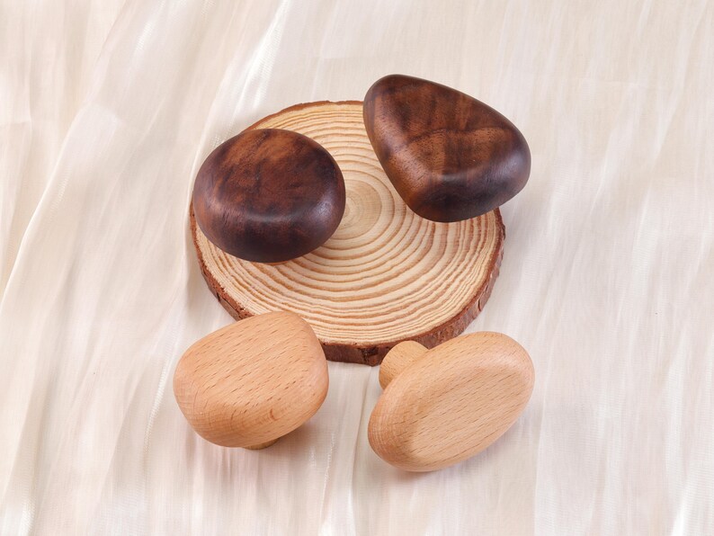 Może przedstawiać: Kolekcja drewnianych gałek do szafek w r&oacute;żnych kształtach i odcieniach drewna. Dwie ciemnobrązowe gałki leżą na drewnianym plastrze, a pod nimi dwie jaśniejsze gałki. Gałki są ułożone na kremowej tkaninie.