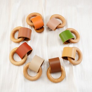 Peut inclure: Une collection d'anneaux de dentition en bois pour b&eacute;b&eacute;s avec des accents en cuir color&eacute;s. Les anneaux sont dispos&eacute;s en cercle, pr&eacute;sentant diff&eacute;rentes couleurs de cuir comme l'orange, le beige, le marron et le vert. Les anneaux en bois sont lisses et de couleur naturelle.