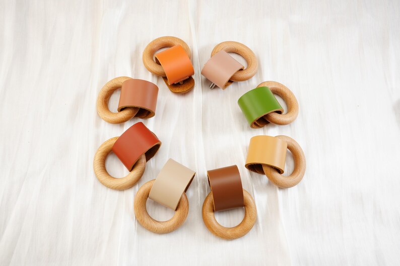 Peut inclure: Une collection d'anneaux de dentition en bois pour b&eacute;b&eacute;s avec des accents en cuir color&eacute;s. Les anneaux sont dispos&eacute;s en cercle, pr&eacute;sentant diff&eacute;rentes couleurs de cuir comme l'orange, le beige, le marron et le vert. Les anneaux en bois sont lisses et de couleur naturelle.