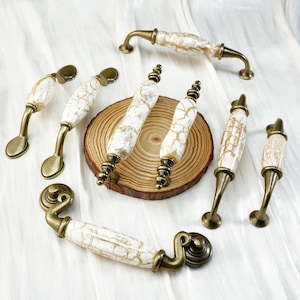 May include: A collection of antique-style cabinet pulls and handles. The hardware features a white and gold crackle finish on the handles, with brass-toned metal accents. Various shapes and sizes are displayed, suitable for furniture restoration or new builds.