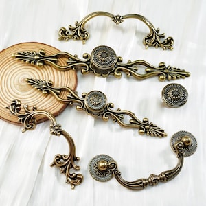 May include: A selection of antique brass cabinet hardware, including handles and knobs. The hardware features ornate floral and scrollwork designs. The pieces are arranged on a white surface with a wooden slice.