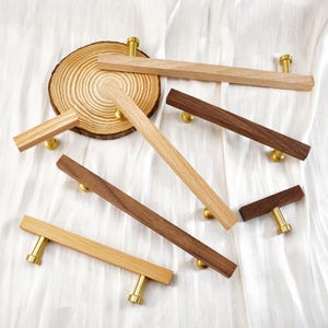 May include: A collection of wooden cabinet pulls in various shades of brown and tan, with brass-colored hardware. The pulls are arranged on a white fabric surface, with a wooden slice in the background.