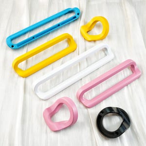 Puede incluir: Un surtido de formas geométricas coloridas en azul, amarillo, blanco y rosa. Las formas incluyen formas rectangulares y circulares, con un anillo negro en la parte inferior. Los objetos están dispuestos sobre una superficie blanca texturizada.