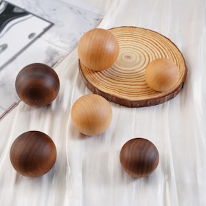 Puede incluir: Esferas de madera en tonos marrones y beige, dispuestas sobre una superficie blanca. Algunas esferas son oscuras, otras claras, y algunas descansan sobre una rodaja de madera. Las esferas son lisas y redondas, sugiriendo una est&eacute;tica natural y artesanal.