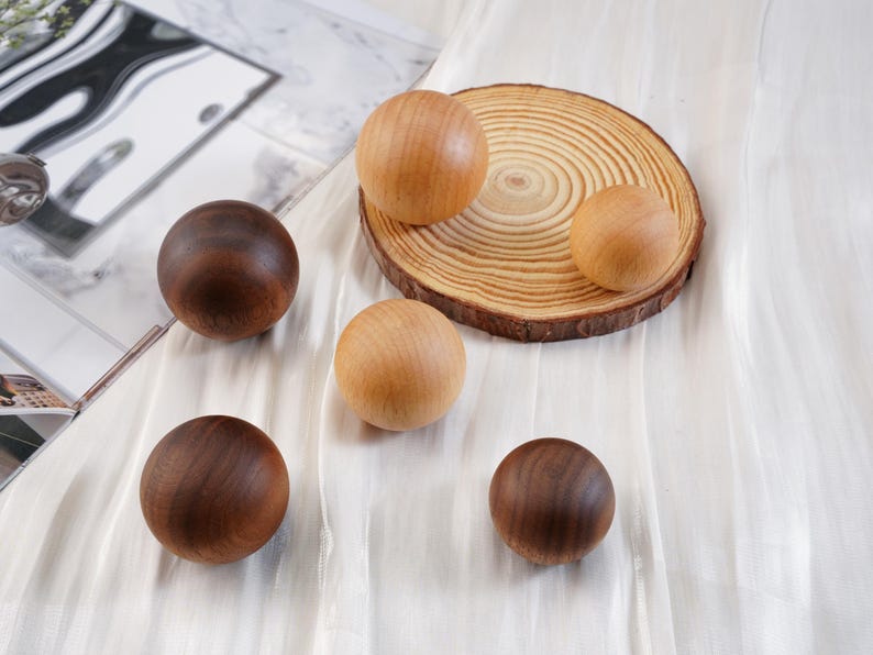 Puede incluir: Esferas de madera en tonos marrones y beige, dispuestas sobre una superficie blanca. Algunas esferas son oscuras, otras claras, y algunas descansan sobre una rodaja de madera. Las esferas son lisas y redondas, sugiriendo una est&eacute;tica natural y artesanal.