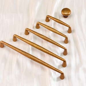 May include: A set of five antique brass cabinet pulls and a matching knob. The pulls are long and slender with curved ends, and the knob is round. The hardware has a warm, aged finish, suggesting a vintage or rustic style.