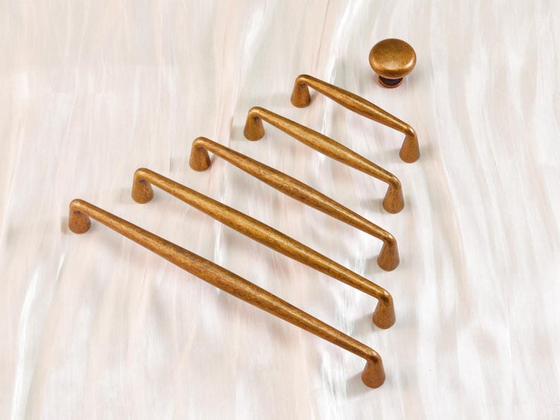 May include: A set of five antique brass cabinet pulls and a matching knob. The pulls are long and slender with curved ends, and the knob is round. The hardware has a warm, aged finish, suggesting a vintage or rustic style.