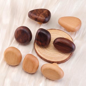 Może przedstawiać: Kolekcja drewnianych gałek do szafek w r&oacute;żnych kształtach i odcieniach brązu i beżu. Niekt&oacute;re gałki są ciemne, inne jasne, a niekt&oacute;re umieszczone na małym kawałku drewna. Gałki ułożone są na białej, teksturowanej powierzchni.