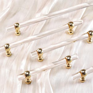 May include: Clear acrylic cabinet pulls with gold-toned hardware. The pulls have a cylindrical shape with gold accents at each end. The hardware is a shiny gold color. The pulls are arranged on a white, textured surface.