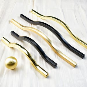 May include: An assortment of cabinet hardware, including curved bar pulls in gold and black finishes, and a gold knob. The hardware is arranged on a white, textured surface, showcasing the variety of designs.