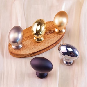 May include: An assortment of decorative cabinet knobs in various metallic finishes. The knobs include silver, gold, and a dark purple. Some knobs are on a wooden oval tray. The knobs are egg-shaped and have a pedestal base.