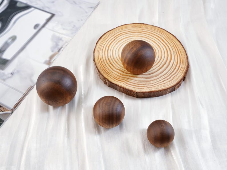 Puede incluir: Cuatro esferas de madera de diferentes tama&ntilde;os est&aacute;n dispuestas sobre una superficie blanca, una de ellas sobre una rodaja de madera circular. Las esferas son de un color marr&oacute;n oscuro intenso, y la rodaja de madera tiene vetas visibles.