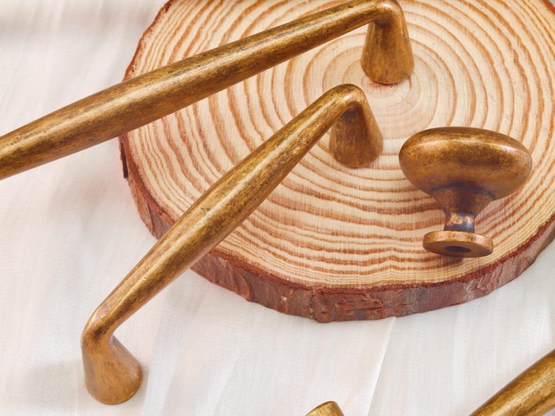 May include: Close-up of antique brass cabinet hardware, including two long pulls and a knob. The hardware has a warm, aged finish and is displayed on a wooden slice with a light-colored background. The pulls are approximately 8 inches long.