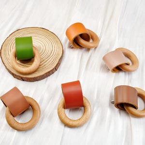 Peut inclure: Anneaux de serviette en bois avec des d&eacute;tails en cuir de diff&eacute;rentes couleurs : vert, orange, beige et marron. Un anneau repose sur une tranche de bois. Les anneaux sont dispos&eacute;s sur un fond blanc textur&eacute;.