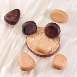 Może przedstawiać: Asortyment drewnianych przedmiot&oacute;w dekoracyjnych w r&oacute;żnych kształtach i odcieniach brązu i beżu. Niekt&oacute;re są ciemne, inne jasne, a jeden spoczywa na małym plasterku drewna. Obiekty ułożone są na białej, teksturowanej powierzchni.