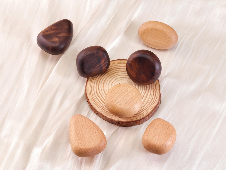 Może przedstawiać: Asortyment drewnianych przedmiot&oacute;w dekoracyjnych w r&oacute;żnych kształtach i odcieniach brązu i beżu. Niekt&oacute;re są ciemne, inne jasne, a jeden spoczywa na małym plasterku drewna. Obiekty ułożone są na białej, teksturowanej powierzchni.