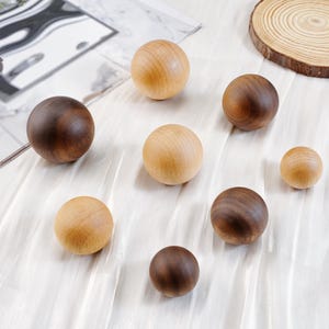 Puede incluir: Una colecci&oacute;n de esferas de madera en varios tama&ntilde;os y tonos de marr&oacute;n y beige. Las esferas est&aacute;n dispuestas sobre una superficie blanca texturizada, con una rodaja de madera natural en el fondo. Las bolas de madera var&iacute;an de marr&oacute;n claro a oscuro.