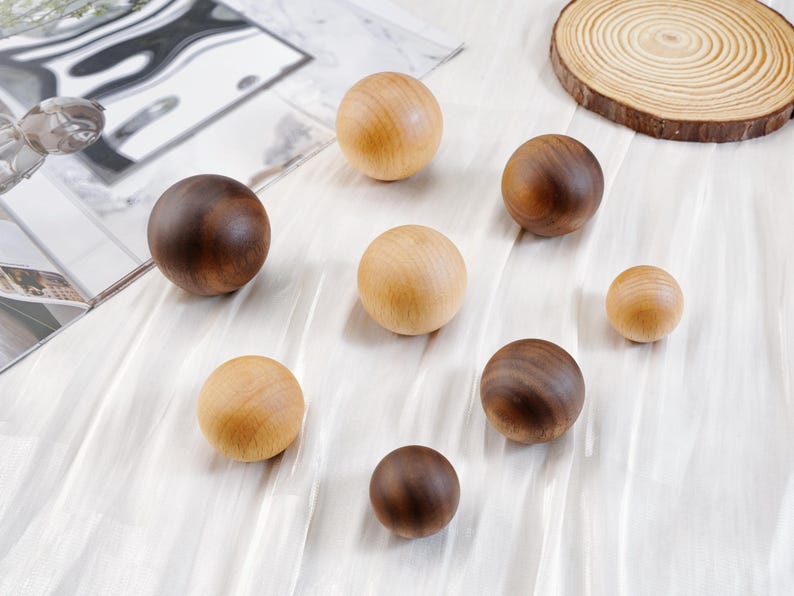 Puede incluir: Una colecci&oacute;n de esferas de madera en varios tama&ntilde;os y tonos de marr&oacute;n y beige. Las esferas est&aacute;n dispuestas sobre una superficie blanca texturizada, con una rodaja de madera natural en el fondo. Las bolas de madera var&iacute;an de marr&oacute;n claro a oscuro.