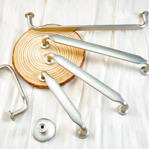 May include: A collection of brushed nickel cabinet hardware, including handles and a knob, arranged on a light-colored fabric and a wooden slice. The hardware features a curved design and a matte finish, suitable for kitchen or bathroom renovations.