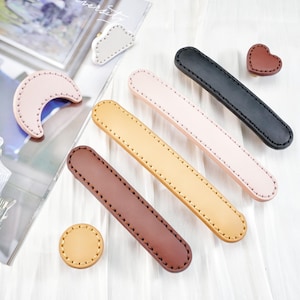 May include: An assortment of leather craft supplies, including handles, a heart, a circle, and a crescent moon shape. The handles are in various colors: black, pink, tan, and brown. All pieces have stitched edges, suggesting a handmade or artisanal quality.