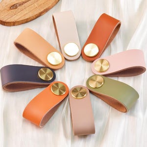 以下が含まれることがあります： オレンジ、ピンク、グリーンなど、さまざまな色合いのカラフルな革製引き出し取っ手のコレクション。各取っ手には真鍮色の円形アクセントが付いています。白い布の表面に円形に配置されています。