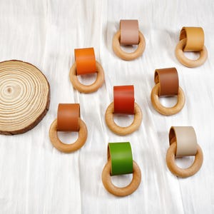 Peut inclure: Anneaux de dentition en bois avec des lani&egrave;res en cuir de diff&eacute;rentes couleurs, dont orange, rouge et vert. Une tranche de bois est &eacute;galement pr&eacute;sente. Les anneaux sont dispos&eacute;s sur une surface blanche.