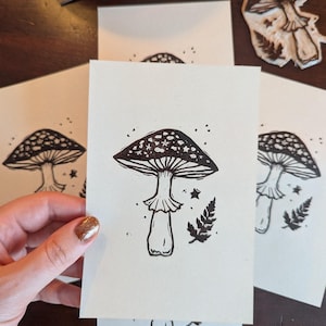 May include: A cream-coloured card held by a hand, displaying a black and white mushroom illustration. The mushroom has a dotted cap, a stem, and is accompanied by stars and fern details. Other cards and a mushroom stamp are visible.