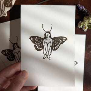 May include: A white rectangular card featuring a black ink block print illustration of a figure with butterfly wings and antennae, sitting with knees tucked towards the chest. The design is centred on the card, showcasing clean lines and high contrast.