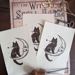 May include: Three cream-coloured prints featuring a black cat sitting on a crescent moon with a face, and a star. The prints are on top of a vintage book page with the text "THE WITCHES SPELL BOOK". A key and dried flowers are also visible.
