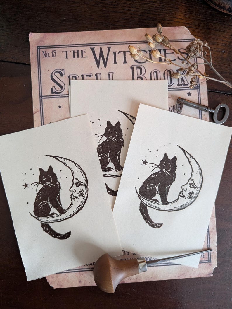 May include: Three cream-coloured prints featuring a black cat sitting on a crescent moon with a face, and a star. The prints are on top of a vintage book page with the text "THE WITCHES SPELL BOOK". A key and dried flowers are also visible.