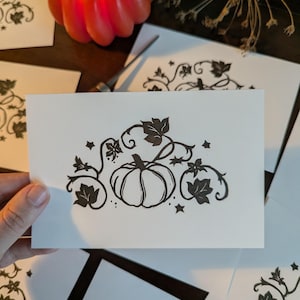 May include: A white rectangular card featuring a black ink illustration of a central pumpkin surrounded by swirling vines, leaves, and small stars. The design is centred on the card, showcasing a botanical theme with clean lines and high contrast.