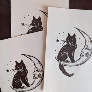 May include: Several cream-coloured art prints depicting a black cat on a crescent moon with a face. The cat gazes at a star. The prints are displayed on a dark wooden surface.