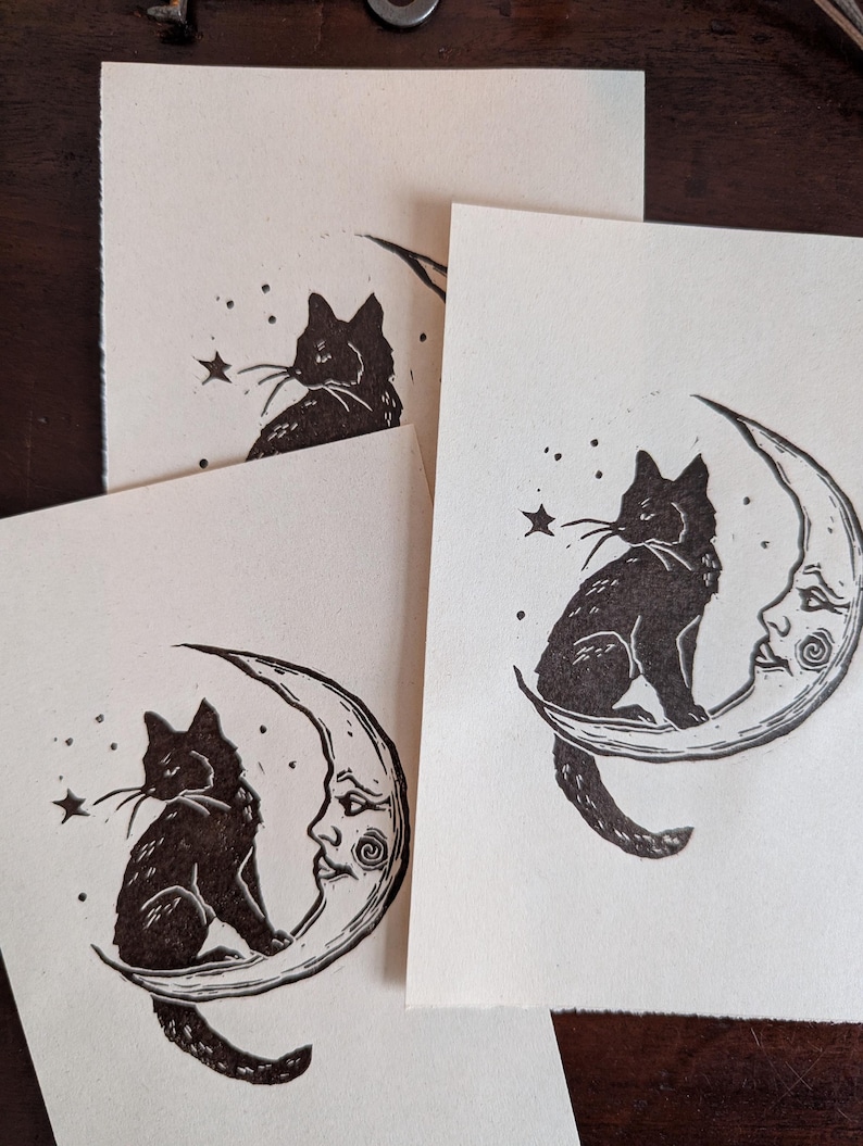 May include: Several cream-coloured art prints depicting a black cat on a crescent moon with a face. The cat gazes at a star. The prints are displayed on a dark wooden surface.