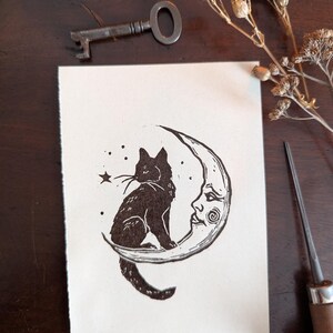 May include: A black and white linocut print depicting a black cat on a crescent moon with a face. The print has a handmade feel. A vintage key and dried flowers are also visible.