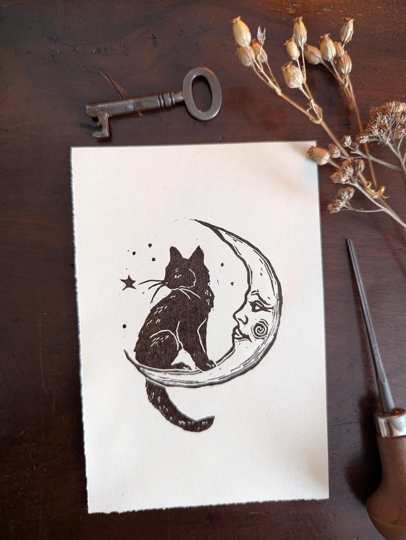 May include: A black and white linocut print depicting a black cat on a crescent moon with a face. The print has a handmade feel. A vintage key and dried flowers are also visible.