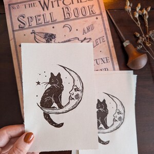 May include: Cream-coloured art prints depicting a black cat on a crescent moon with a face. The prints are next to a vintage book titled "The Witches Spell Book" and a wood carving tool.