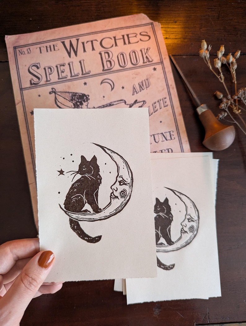 May include: Cream-coloured art prints depicting a black cat on a crescent moon with a face. The prints are next to a vintage book titled "The Witches Spell Book" and a wood carving tool.