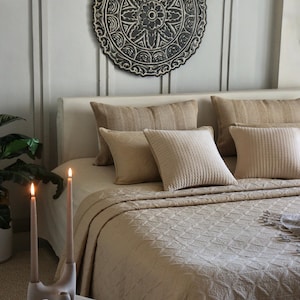 May include: A bedroom interior with a bed dressed in beige bedding and cushions. A decorative circular wall hanging in black and white is above the bed. Lit candles and a book are on a side table.