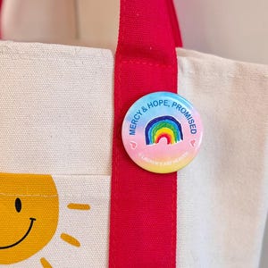 Può includere: Una borsa tote color crema con cinghie rosse brillanti e una tasca frontale con una grafica di sole sorridente e le parole "Love is". Una spilla rotonda con un arcobaleno e le parole "Mercy & Hope, Promised" è attaccata alla cinghia.