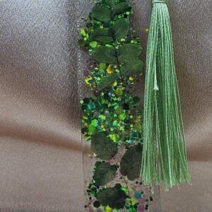 May include: Clear resin bookmark with embedded green leaves, glitter, and a green tassel. The bookmark has a rectangular shape with rounded edges. The glitter includes shades of green, yellow, and purple.