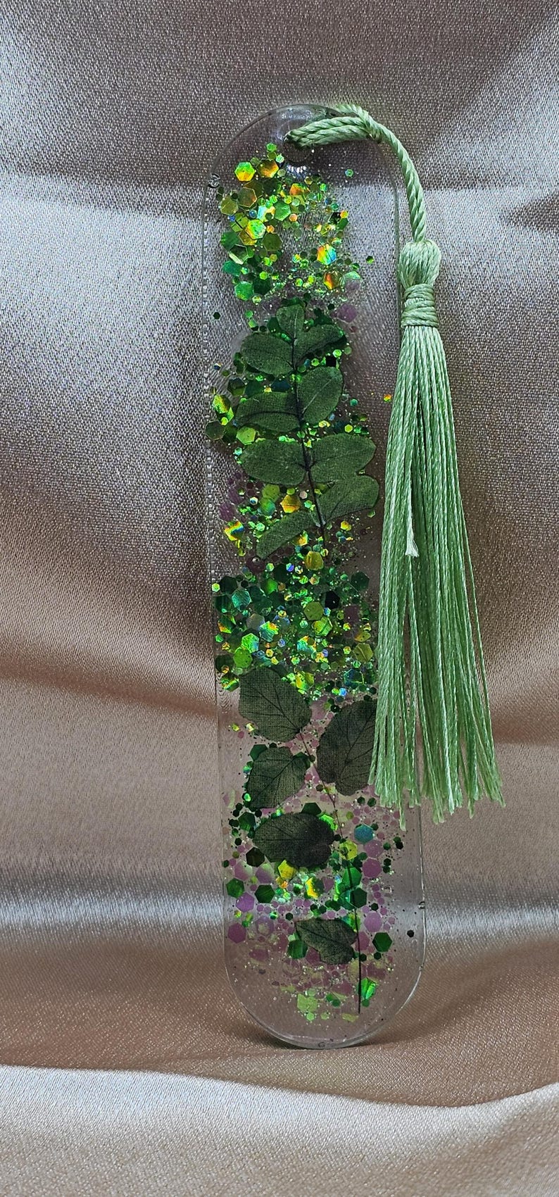 May include: Clear resin bookmark with embedded green leaves, glitter, and a green tassel. The bookmark has a rectangular shape with rounded edges. The glitter includes shades of green, yellow, and purple.