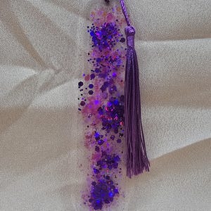 May include: A clear resin bookmark with embedded purple glitter and a matching purple tassel. The bookmark has a rounded bottom and a braided cord attached to the tassel. The glitter is a mix of shapes and sizes.