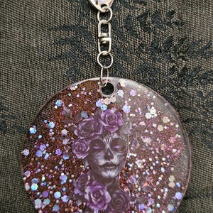 May include: A round keychain featuring a Day of the Dead sugar skull design. The image of the skull is surrounded by purple roses and glitter. The keychain has a silver clasp and chain.
