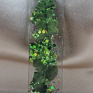 May include: A clear resin bookmark with a green leaf and glitter design. The bookmark features a sprig of leaves and green, yellow, and purple glitter. A silver-colored cord is attached at the top.